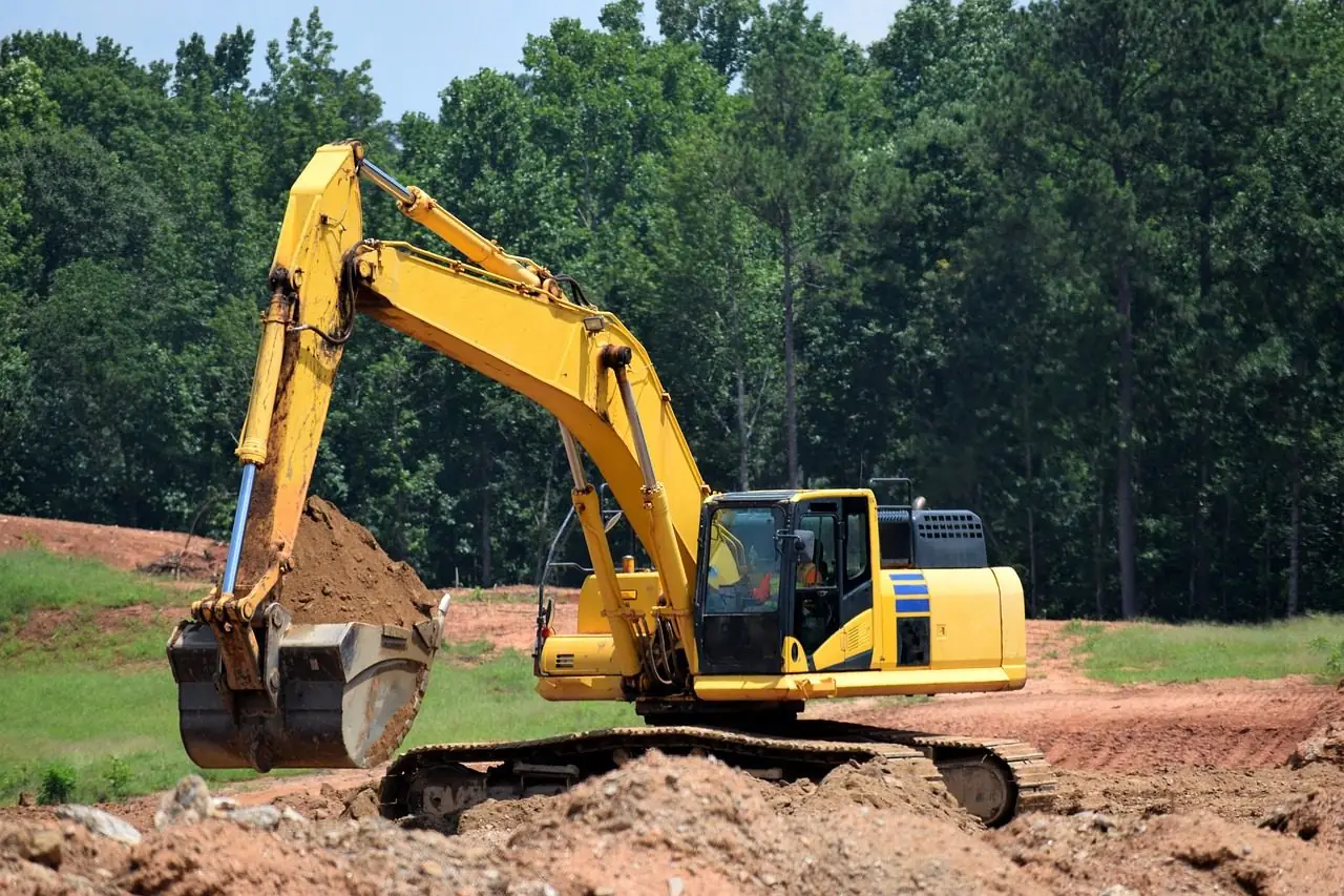 A yellow excavator digging red-brown soil on a cleared site with a pine woods behind
